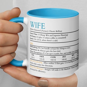 May include: A white ceramic mug with a blue rim and handle. The mug features a humorous character description for "WIFE" with stats and abilities. The text reads: "Wife, Man Humanoid (Partner), Chaotic Brilliant. Armour Class 18 (Sharp Wit and Superior Planning). Hit Points 85 (plus 15 when coffee is consumed). Speed 35 ft. (50 ft. when there's a sale). Multitasking +8: Can handle at least five things at once. Negotiation +6: Always gets the best deal. Divine Perception +7: Can spot crumbs, lies, and trouble from 30 ft. away. STR 12(+1), DEX 14(+2), CON 16(+3), INT 18(+4), WIS 14(+2). Art of Leadership: Allies within 15 feet gain +2 to intelligence and wisdom checks while under her direction. Money De Manager: Assigns 1d4 tasks to the Husband. Tasks are prioritised for efficiency."