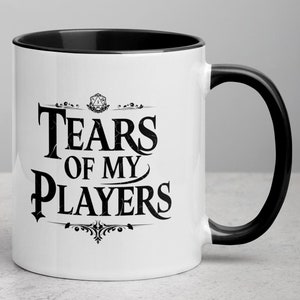 Könnte beinhalten: Weiße Keramik-Kaffeetasse mit schwarzem Rand und Henkel. Die Tasse zeigt eine schwarze Grafik eines Dungeons and Dragons-Würfels mit dem Text "Tears of My Players" in einer dekorativen Schrift.