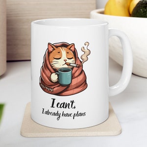 May include: White ceramic mug with a cartoon cat wrapped in a red blanket, holding a cup of coffee and smoking a cigarette. The text on the mug reads "I can't, I already have plans."