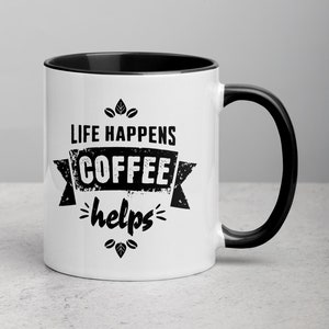 Peut inclure: Une tasse à café en céramique blanche avec un bord et une poignée noirs. La tasse présente un motif noir qui dit "Life Happens Coffee Helps" avec une bannière et des accents de grains de café.