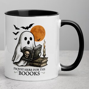 Op de afbeelding: Witte keramische mok met een zwarte binnenkant en handvat. De mok heeft een Halloween-thema illustratie van een geest die een boek leest met een schedel op de kaft, vleermuizen en een volle oranje maan. Tekst op de mok luidt "I'M JUST HERE FOR THE BOOOOKS."
