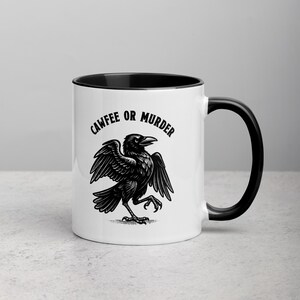 May include: White ceramic mug with a black interior and handle. The mug features a black illustration of a crow with outstretched wings and the text "CAWFEE OR MURDER" in a black, stylized font.