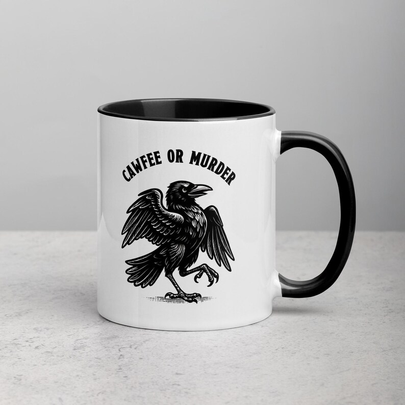 May include: White ceramic mug with a black interior and handle. The mug features a black illustration of a crow with outstretched wings and the text "CAWFEE OR MURDER" in a black, stylized font.