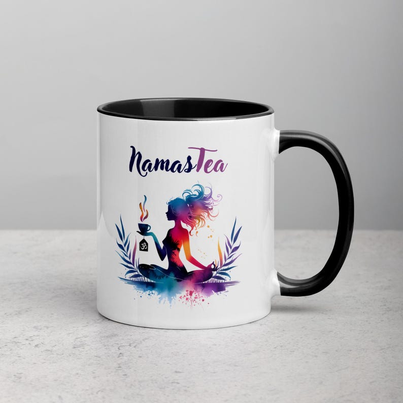 K&ouml;nnte beinhalten: Wei&szlig;e Keramiktasse mit schwarzem Rand und Henkel. Die Tasse zeigt eine farbenfrohe Illustration einer Frau in einer Yoga-Pose, die eine Tasse Tee h&auml;lt. Der Text "Namastea" ist in Schwarz auf die Tasse gedruckt.