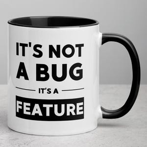 Peut inclure: Mug en céramique blanche avec intérieur et anse noirs. Le mug affiche le texte "IT'S NOT A BUG IT'S A FEATURE" en lettres noires grasses. Un design humoristique pour le café ou le thé.