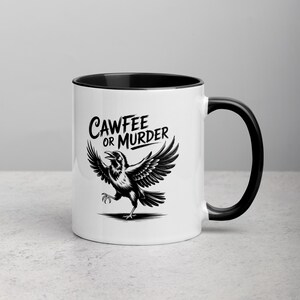 May include: White ceramic mug with a black interior and handle. The mug features a black and white illustration of a crow with outstretched wings, and the text "CAWFEE OR MURDER" in a stylized font.
