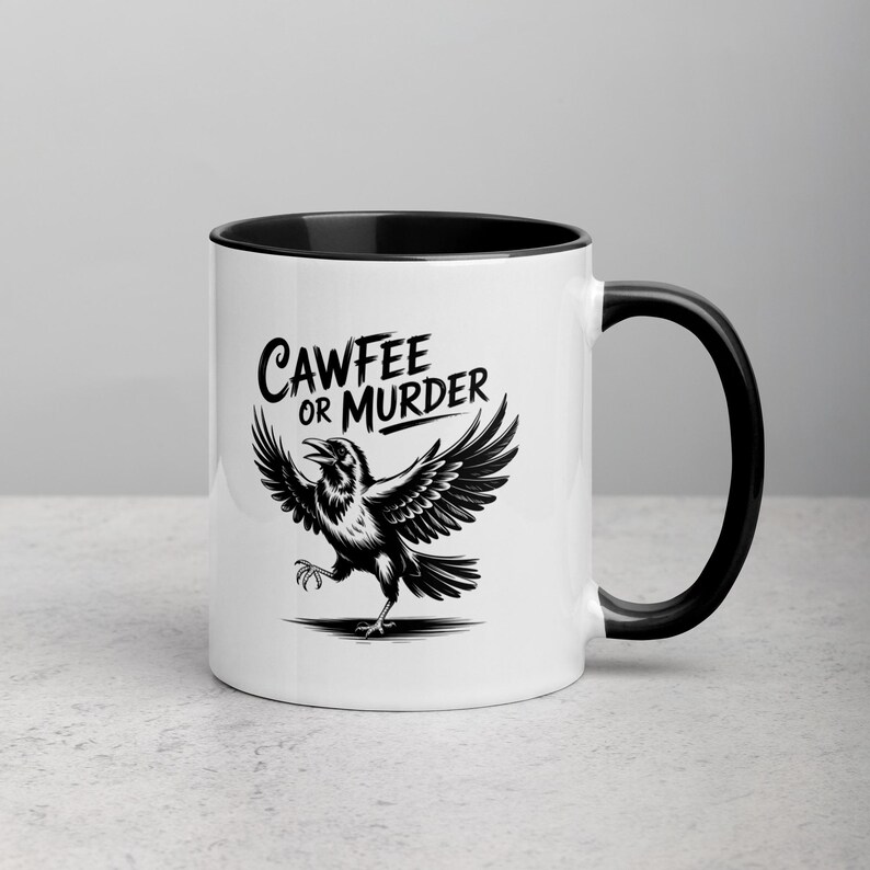 May include: White ceramic mug with a black interior and handle. The mug features a black and white illustration of a crow with outstretched wings, and the text "CAWFEE OR MURDER" in a stylized font.