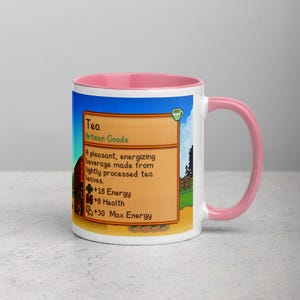 May include: White ceramic mug with a pink interior and handle. The mug features a colorful graphic from a video game, including text that reads "Tea" and details about its effects. The design is set against a blue sky background.