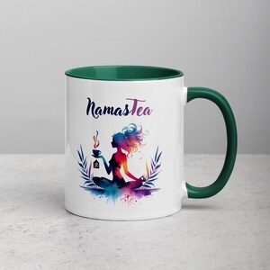 K&ouml;nnte beinhalten: Wei&szlig;e Keramiktasse mit gr&uuml;nem Rand und Henkel. Die Tasse zeigt eine farbenfrohe Illustration einer meditierenden Frau mit einer Tasse Tee und dem Text "Namastea".