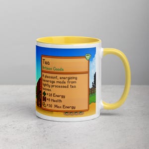 May include: A white ceramic mug with a yellow interior and handle. The mug features a colorful graphic from a video game, including text that reads "Tea" and details about the beverage's effects. The design is set against a blue sky background.