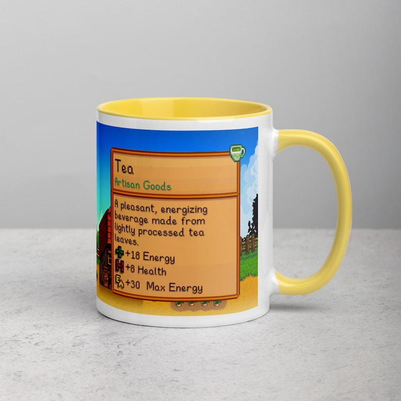 May include: A white ceramic mug with a yellow interior and handle. The mug features a colorful graphic from a video game, including text that reads "Tea" and details about the beverage's effects. The design is set against a blue sky background.