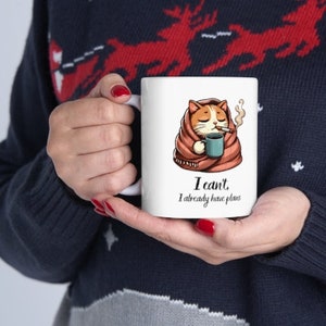 May include: White ceramic mug with a cartoon illustration of a cat wrapped in a blanket, holding a cup of coffee and smoking a cigarette. The text on the mug reads "I can't. I already have plans."