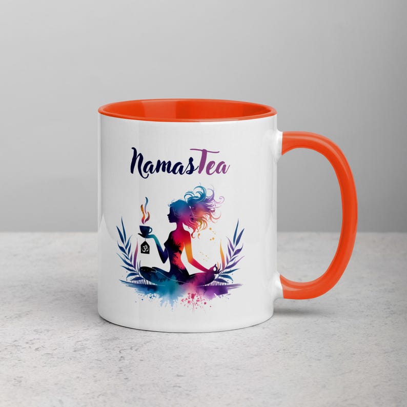 K&ouml;nnte beinhalten: Wei&szlig;e Keramiktasse mit orangefarbenem Rand und Henkel. Die Tasse zeigt eine farbenfrohe Illustration einer Frau, die in einer Yoga-Pose sitzt und eine Tasse Tee h&auml;lt. Der Text "Namastea" ist in Schwarz auf die Tasse gedruckt.