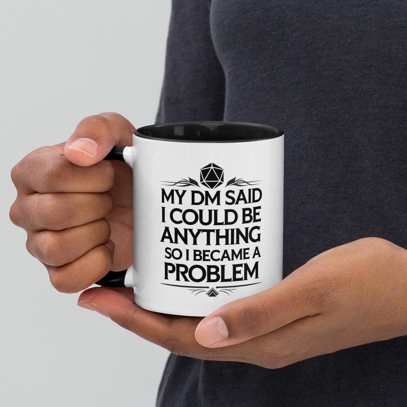 May include: White ceramic mug with a black interior and handle. The mug features the text "MY DM SAID I COULD BE ANYTHING SO I BECAME A PROBLEM" in a black, bold font, with a geometric design above and below the text. The mug is held by a person.