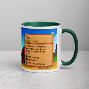 May include: White ceramic mug with a green handle and interior. The mug features a colorful graphic from a video game, including the word "Tea" and details about the beverage's properties. The mug is a standard size for hot beverages.