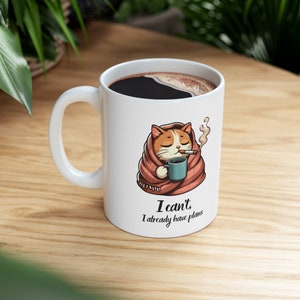 May include: White ceramic mug with a cartoon cat wrapped in a blanket, holding a cup of coffee and smoking a cigarette. The text on the mug reads "I can't, I already have plans."