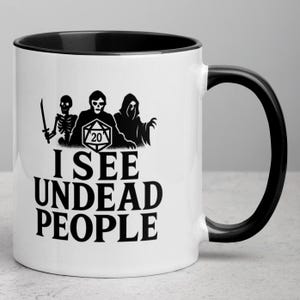 May include: White ceramic mug with a black interior and handle. The mug features a black graphic of a skeleton, a figure with a dice, and a hooded figure. The text "I SEE UNDEAD PEOPLE" is printed below the graphic.