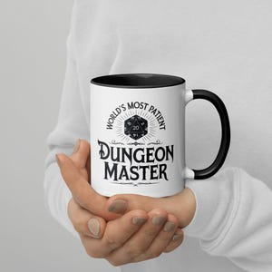 May include: White ceramic mug with a black rim and handle. The mug has a black graphic that says "World's Most Patient Dungeon Master" with a black and white 20-sided die graphic.