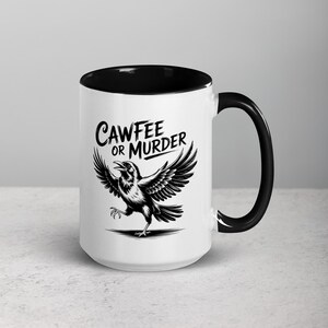 May include: White ceramic coffee mug with a black interior and handle. The mug features a black and white illustration of a crow with outstretched wings and the text "CAWFEE OR MURDER" in a stylized font.