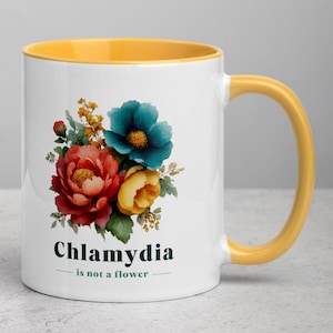 May include: White ceramic mug with a yellow interior and handle. The mug features a floral design with red, blue, and yellow flowers. The text "Chlamydia is not a flower" is printed below the flowers.