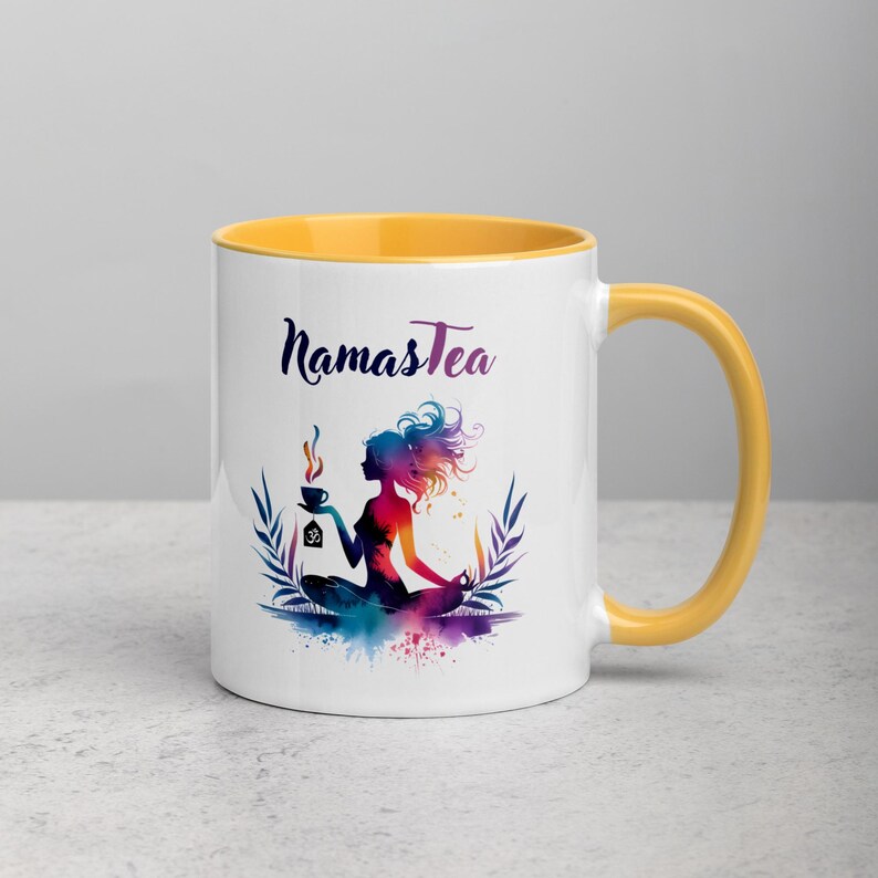 K&ouml;nnte beinhalten: Wei&szlig;e Keramiktasse mit gelbem Rand und Henkel. Die Tasse zeigt eine farbenfrohe Illustration einer Frau in einer Yoga-Pose, die eine Teetasse h&auml;lt. Der Text "Namastea" ist in Schwarz auf die Tasse gedruckt.