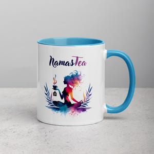 K&ouml;nnte beinhalten: Wei&szlig;e Keramiktasse mit hellblauem Rand und Henkel. Die Tasse zeigt eine farbenfrohe Illustration einer Frau in einer Yoga-Pose, die eine Tasse Tee h&auml;lt. Der Text "Namastea" ist in Schwarz auf die Tasse gedruckt.