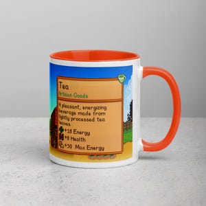 May include: White ceramic mug with an orange interior and handle. The mug features a colorful graphic from a video game, including text that reads "Tea" and details about the beverage. The design is set against a blue sky and green landscape.