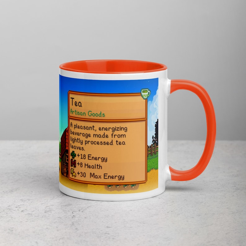May include: White ceramic mug with an orange interior and handle. The mug features a colorful graphic from a video game, including text that reads "Tea" and details about the beverage. The design is set against a blue sky and green landscape.