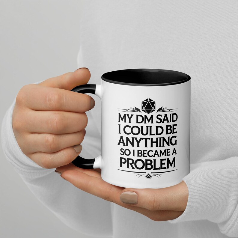 May include: White ceramic mug with a black interior and handle. The mug features the text "MY DM SAID I COULD BE ANYTHING SO I BECAME A PROBLEM" in black, along with a geometric design. The mug is held by a person.