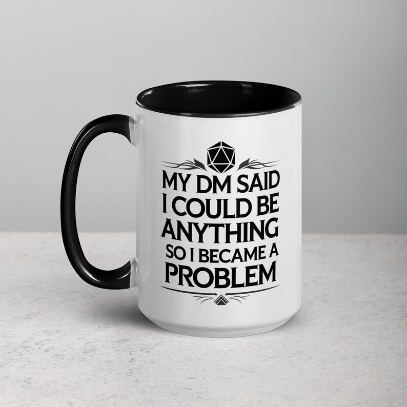 May include: White ceramic mug with a black interior and handle. The mug features the text "MY DM SAID I COULD BE ANYTHING SO I BECAME A PROBLEM" in black, along with a geometric design. The mug is designed for fans of tabletop role-playing games.