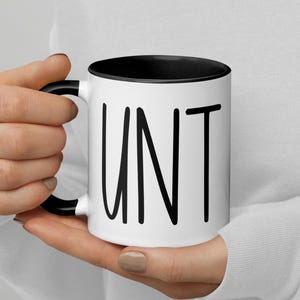 Peut inclure: Une tasse en céramique blanche avec un bord et une poignée noirs. La tasse a le mot "UNT" écrit en noir sur le devant.