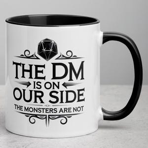 Op de afbeelding: Een witte keramische mok met een zwarte rand en handvat. De mok heeft een zwart-witte afbeelding van een 20-zijdige dobbelsteen met de tekst "The DM is on our side. The monsters are not."