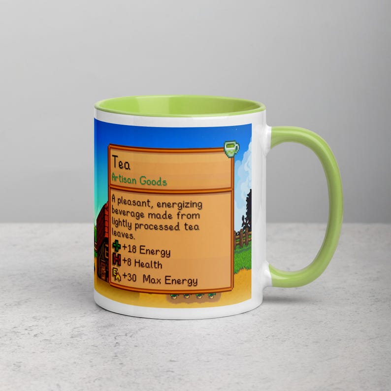 May include: White ceramic mug with a green interior and handle. The mug features a colorful graphic from a video game, including text that reads "Tea" and details about its effects. The design includes a blue sky, a farm scene, and a pixelated style.