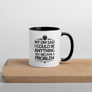 May include: White ceramic mug with a black interior and handle. The mug features the text "MY DM SAID I COULD BE ANYTHING SO I BECAME A PROBLEM" in black, with a geometric design above the text. The mug is on a wooden surface.