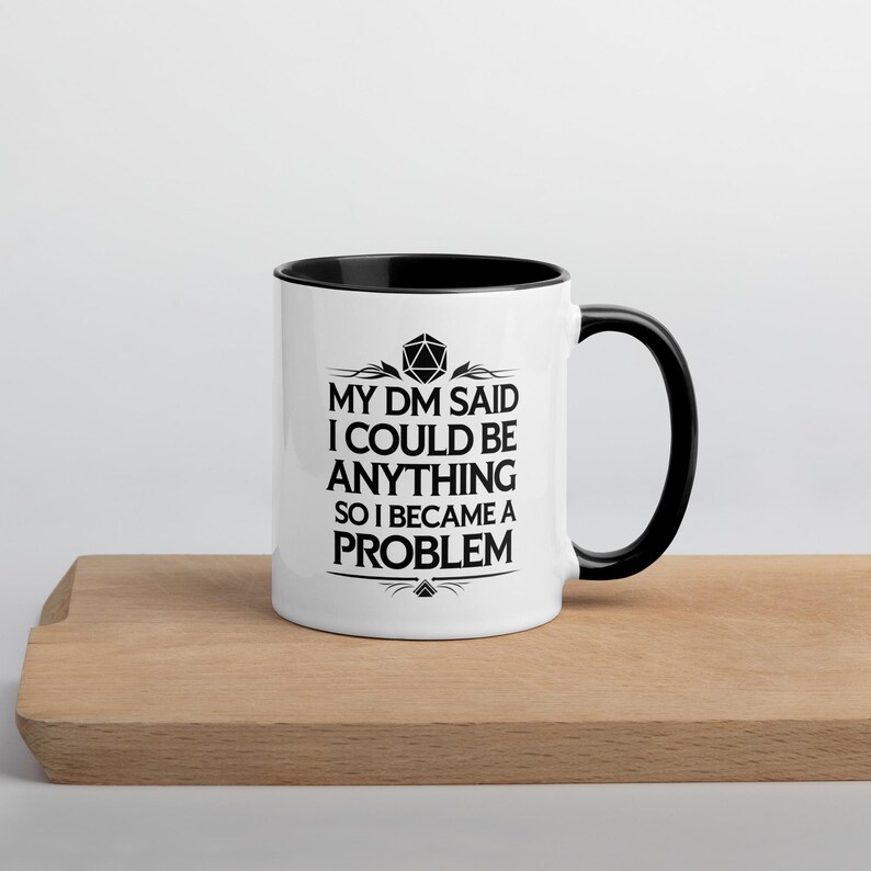 May include: White ceramic mug with a black interior and handle. The mug features the text "MY DM SAID I COULD BE ANYTHING SO I BECAME A PROBLEM" in black, with a geometric design above the text. The mug is on a wooden surface.