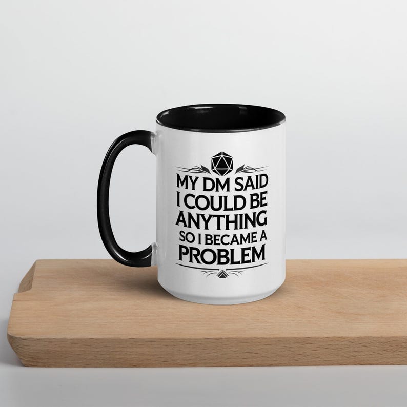 May include: White ceramic mug with a black interior and handle. The mug features the text "MY DM SAID I COULD BE ANYTHING SO I BECAME A PROBLEM" in black, along with a graphic of a 20-sided die. The mug is on a wooden surface.