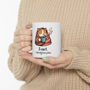 May include: White ceramic mug with a cartoon cat wrapped in a blanket, holding a cup of coffee and smoking a cigarette. The text on the mug reads "I can't, I already have plans."