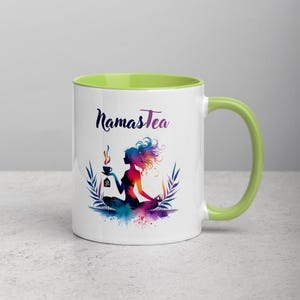 K&ouml;nnte beinhalten: Wei&szlig;e Keramiktasse mit gr&uuml;nem Rand und Henkel. Die Tasse zeigt eine farbenfrohe Illustration einer Frau, die mit einer Tasse Tee meditiert, und den Text "Namastea".