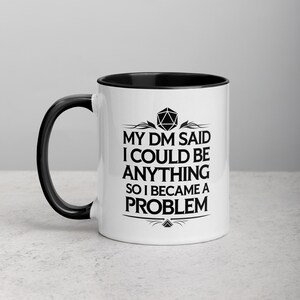 May include: White ceramic mug with a black interior and handle. The mug features the text "MY DM SAID I COULD BE ANYTHING SO I BECAME A PROBLEM" in black, along with a geometric design. A fun gift for a Dungeons and Dragons player.