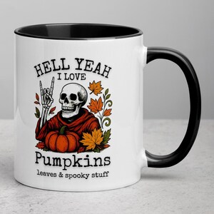 May include: White ceramic mug with a black interior and handle. The mug features a Halloween-themed design with a skeleton wearing a red hoodie, a pumpkin, and the text "HELL YEAH I LOVE PUMPKINS leaves & spooky stuff."