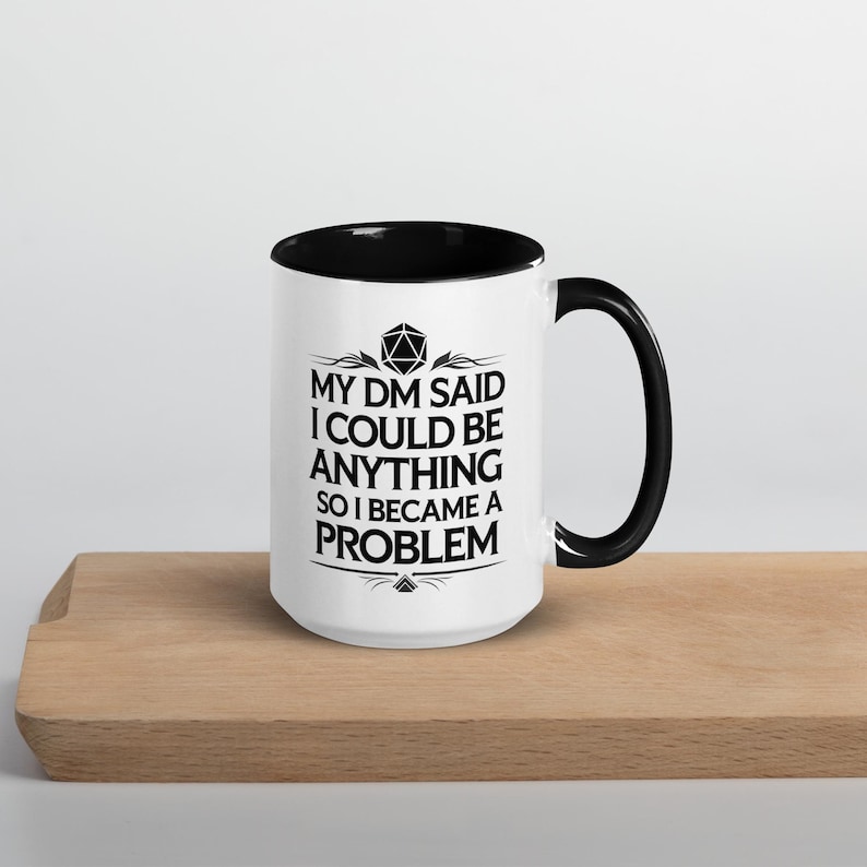 May include: White ceramic mug with a black interior and handle. The mug features the text "MY DM SAID I COULD BE ANYTHING SO I BECAME A PROBLEM" in black, with decorative elements above and below the text. The mug is on a wooden surface.