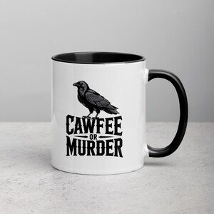 May include: White ceramic mug with a black interior and handle. The mug features a black illustration of a raven perched above the words "CAWFEE OR MURDER" in a bold, stylized font. The mug is on a light gray surface.