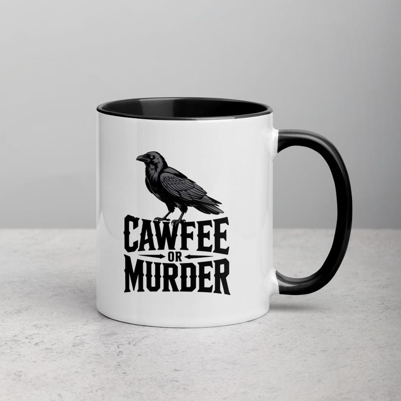 May include: White ceramic mug with a black interior and handle. The mug features a black illustration of a raven perched above the words "CAWFEE OR MURDER" in a bold, stylized font. The mug is on a light gray surface.