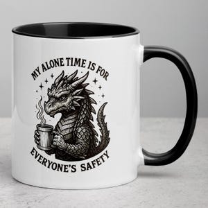 May include: White ceramic mug with a black interior and handle. The mug features a detailed illustration of a dragon holding a mug of coffee, with the text "MY ALONE TIME IS FOR EVERYONE'S SAFETY" in a circular design.