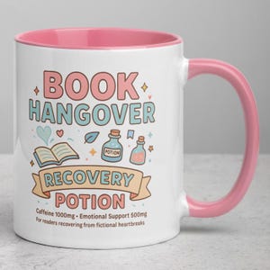 Könnte beinhalten: Weißer Keramikbecher mit rosa Innenraum und Henkel. Der Becher zeigt den Text "BOOK HANGOVER RECOVERY POTION" mit Illustrationen eines Buches und von Zaubertränken. Der Text darunter lautet "Caffeine 1000mg - Emotional Support 500mg".