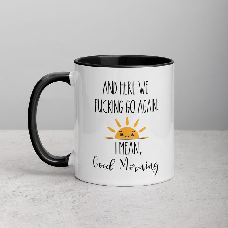 Pu&ograve; includere: Tazza da caff&egrave; in ceramica bianca con bordo e manico neri. La tazza presenta una grafica in bianco e nero di un sole sorridente con il testo "And here we fucking go again. I mean, Good Morning."
