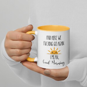 Pu&ograve; includere: Tazza da caff&egrave; in ceramica bianca con un bordo giallo. La tazza ha una grafica in bianco e nero di un sole sorridente con il testo "And here we fucking go again. I mean, Good Morning".