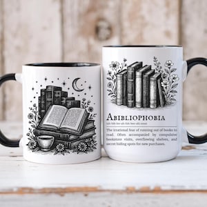 Könnte beinhalten: Zwei weiße Keramikbecher mit schwarzen Rändern und Griffen. Ein Becher zeigt einen Stapel Bücher, ein offenes Buch, eine Tasse, einen Mond und Sterne. Der andere Becher hat das Wort "ABIBLIOPHOBIA" und eine Definition mit einem Stapel Bücher.