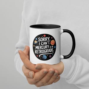 Peut inclure: Une tasse en c&eacute;ramique blanche avec un bord et une poign&eacute;e noirs. La tasse pr&eacute;sente un cercle noir avec un motif spatial et le texte "D&eacute;sol&eacute;, je ne peux pas. Mercure est r&eacute;trograde."
