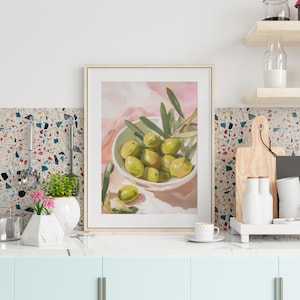May include: A framed painting depicts a still life of green olives in a white bowl with olive branches. The bowl sits on a pink cloth, with a light wood frame. The painting is displayed in a kitchen setting.